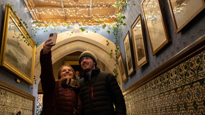 A young couple huddle together in the corridor at Cragside House to take a selfie under sparkling warm white lights draped across the ceiling.